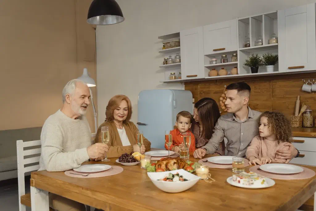 Trois générations réunies autour d'une table de cuisine en bois : un couple de grands-parents, leurs enfants adultes et deux jeunes enfants