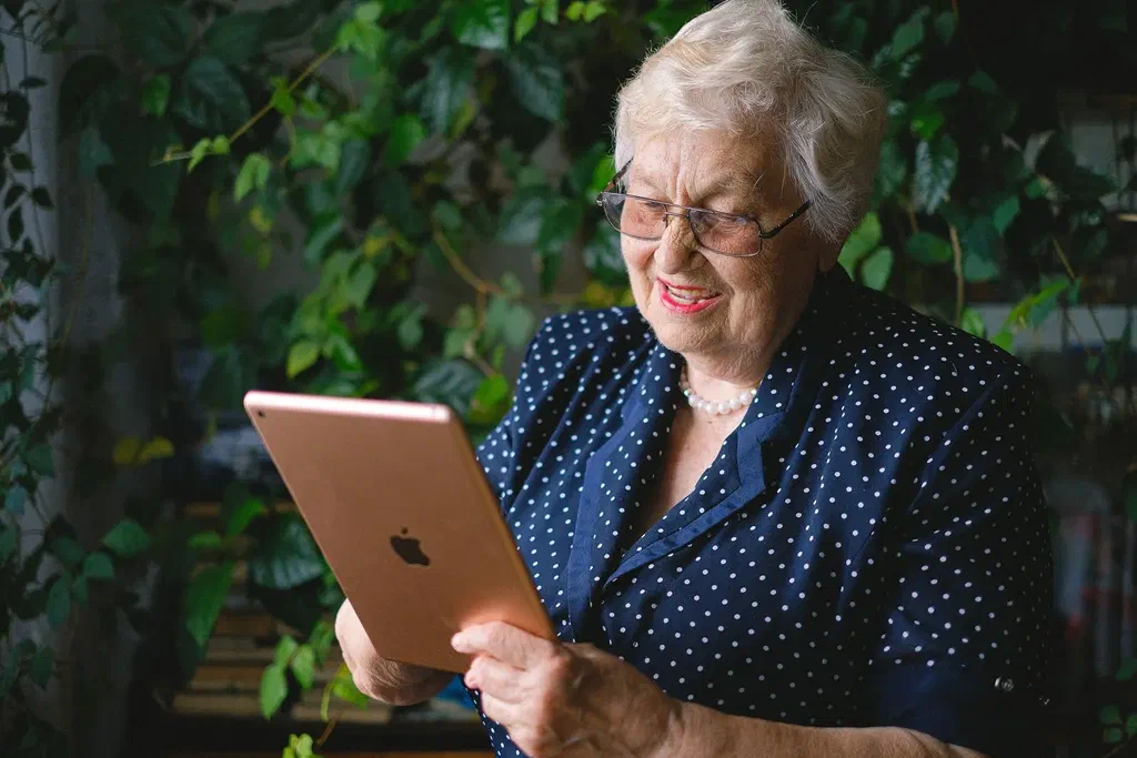 Une femme âgée aux cheveux blancs sourit en tenant une tablette devant elle, en appel vidéo, devant des plantes vertes