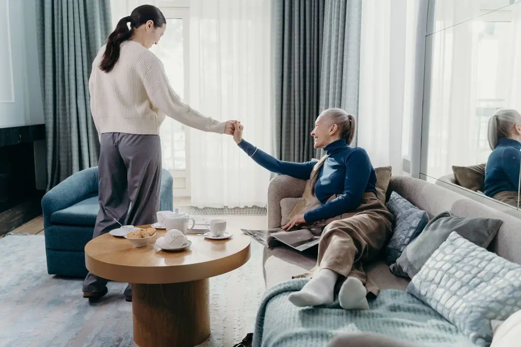 Une femme aide sa mère âgée à se lever d'un canapé dans un salon lumineux