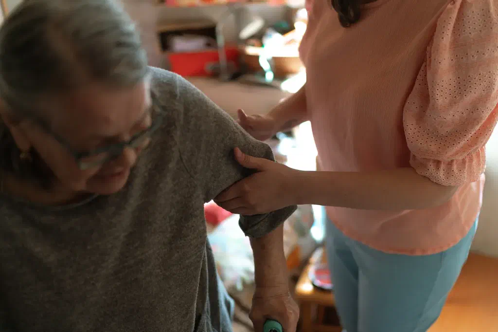 Une femme aide une personne âgée appuyée sur une canne à se redresser dans son salon