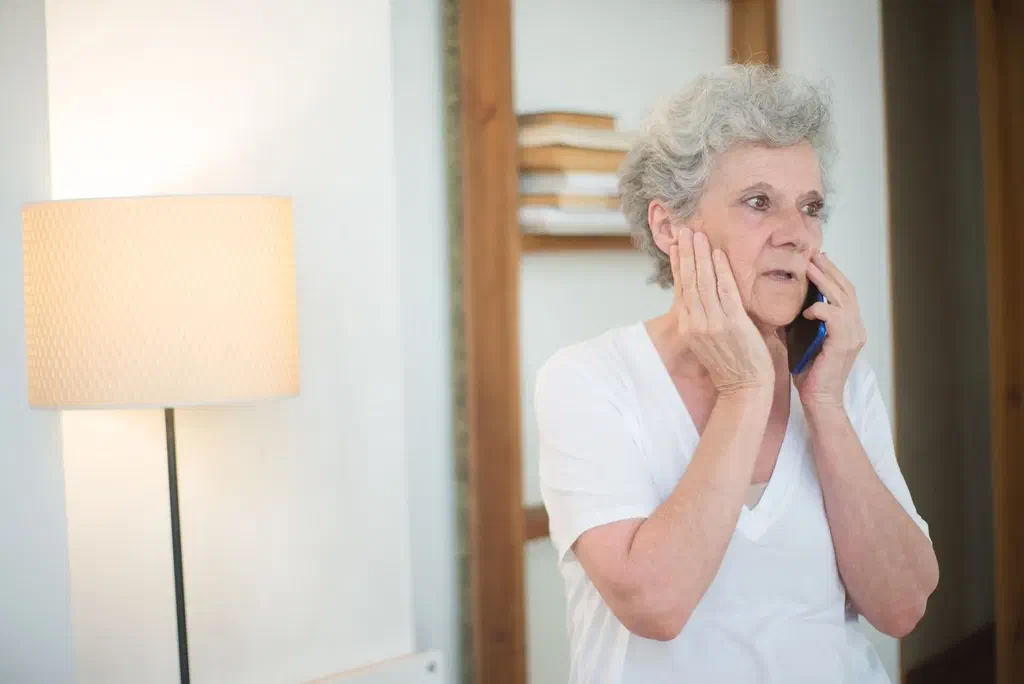 Une femme âgée aux cheveux gris, au téléphone dans son salon, la main sur la joue, l'air inquiet