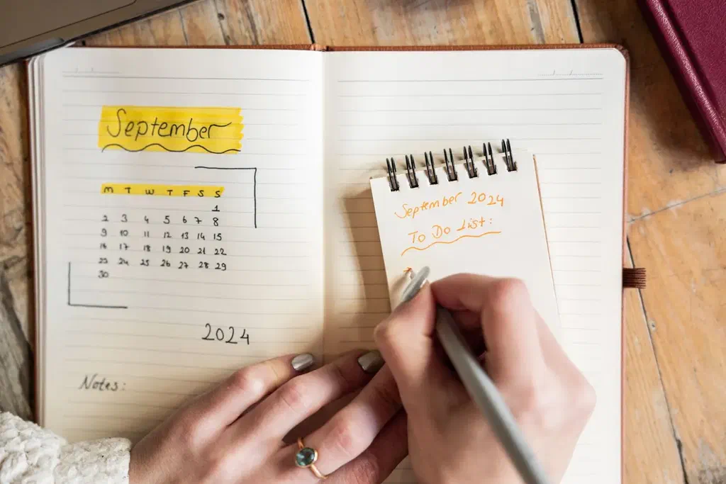 Une main écrit une liste de tâches dans un carnet ouvert sur une table en bois