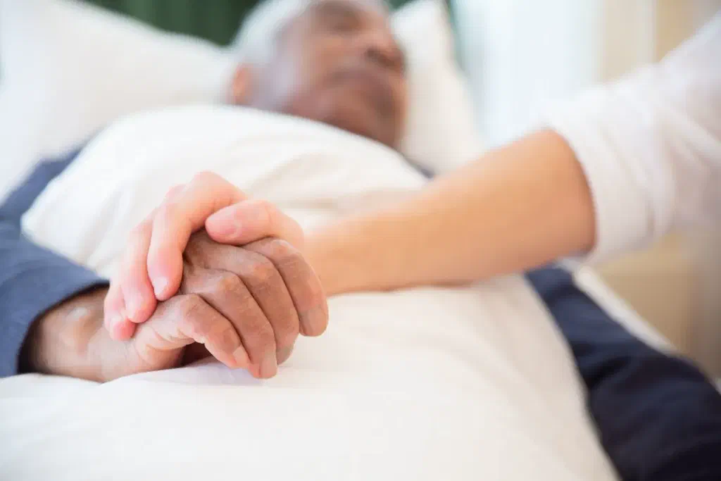 Une main tient doucement celle d'un homme âgé allongé dans un lit, sous une couette blanche