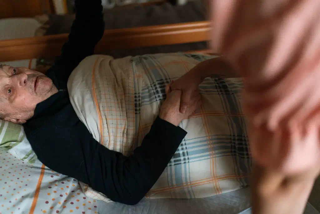 Une femme âgée allongée dans son lit, chez elle, tient la main d'une aidante penchée vers elle