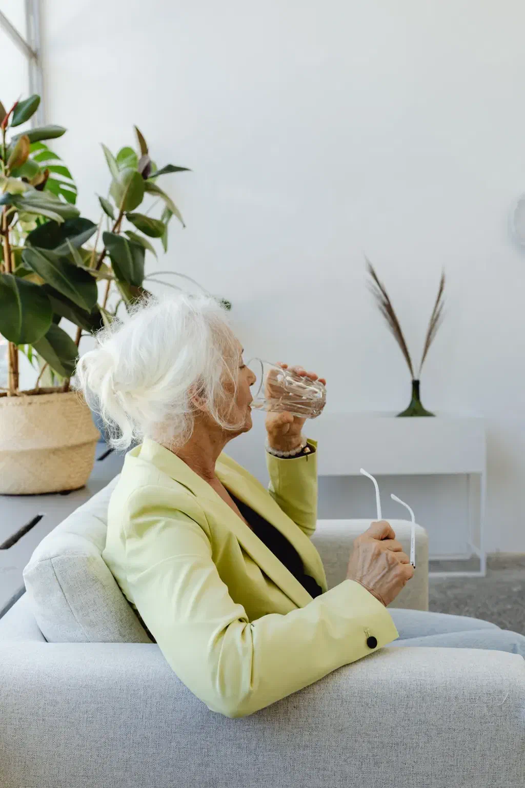 Une femme âgée assise dans un fauteuil clair boit un verre d'eau