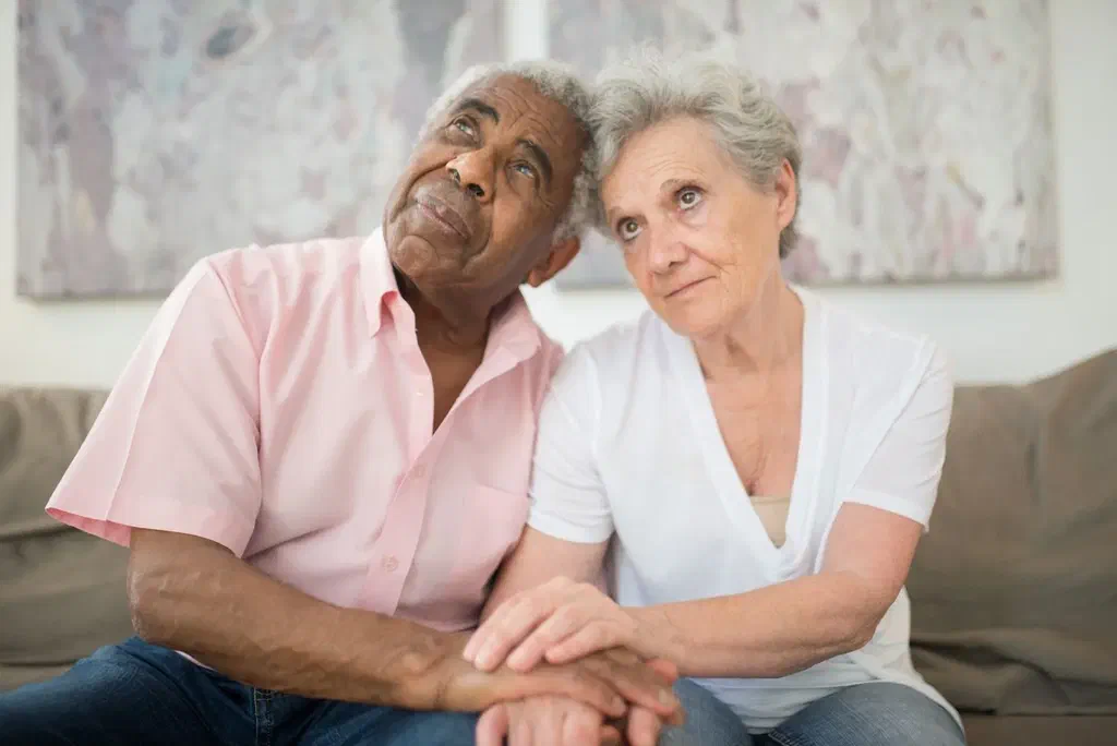 Un couple âgé assis côte à côte sur un canapé, les mains jointes, le regard levé