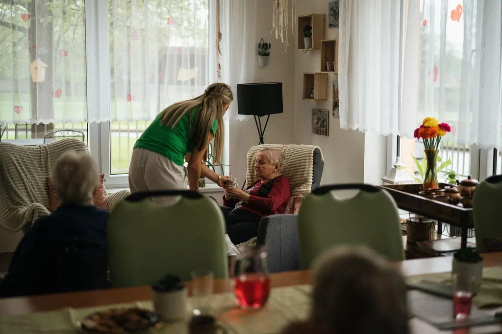 La salle commune lumineuse d'un établissement, où une soignante en tee-shirt vert se penche vers une résidente assise dans un fauteuil