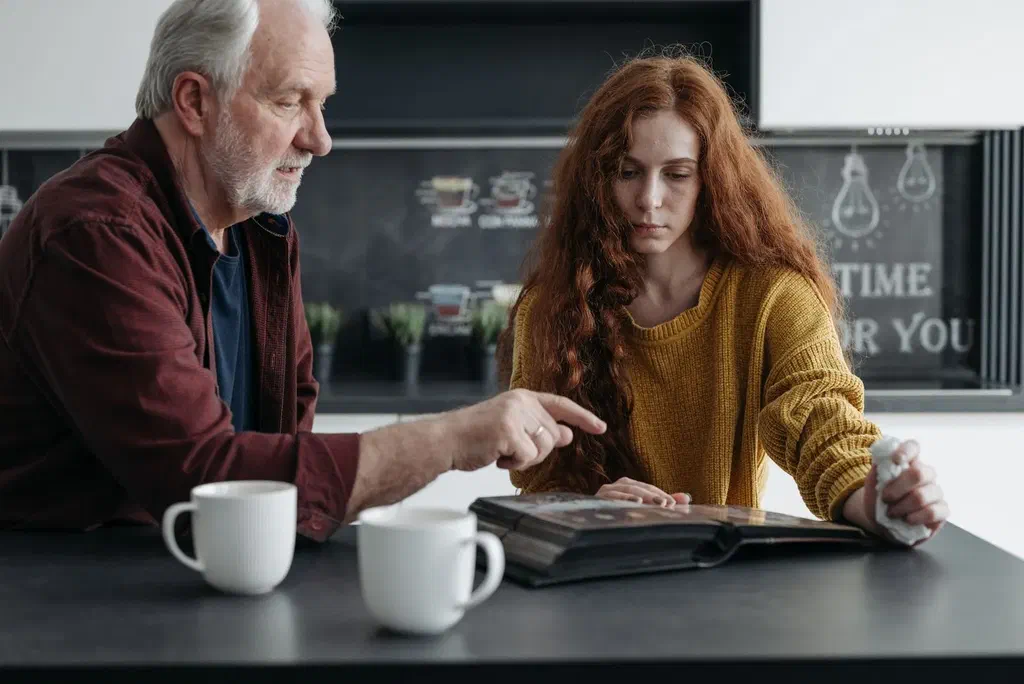 Un homme âgé aux cheveux blancs et une jeune femme feuillettent un album photo à une table de cuisine, deux tasses posées devant eux