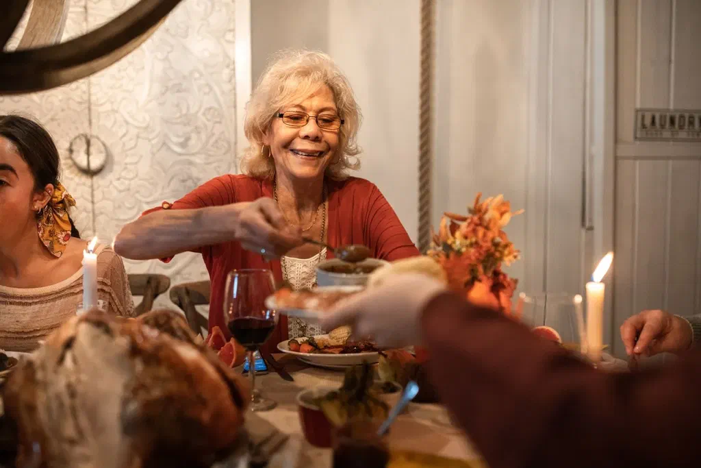 Une femme âgée aux lunettes, souriante, se sert à table lors d'un repas de famille éclairé à la bougie