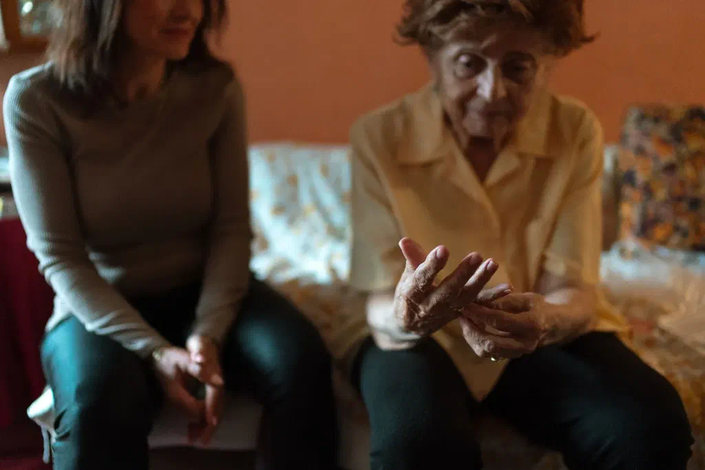 Une femme âgée en chemisier jaune parle avec les mains, assise sur un canapé à côté d'une femme plus jeune qui l'écoute