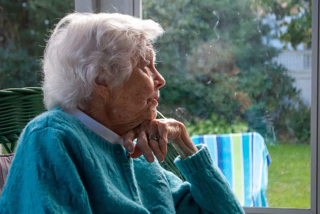 Une femme âgée aux cheveux blancs, en gilet bleu, le menton posé sur la main, regarde par une fenêtre donnant sur un jardin