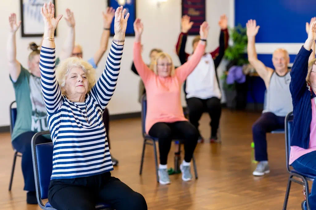 Dans une salle municipale, un groupe de retraités assis sur des chaises lève les bras au-dessus de la tête pendant un cours collectif
