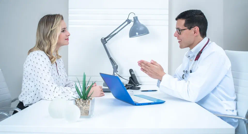 Une femme blonde en chemisier échange avec un médecin en blouse blanche, assis de part et d'autre d'un bureau clair