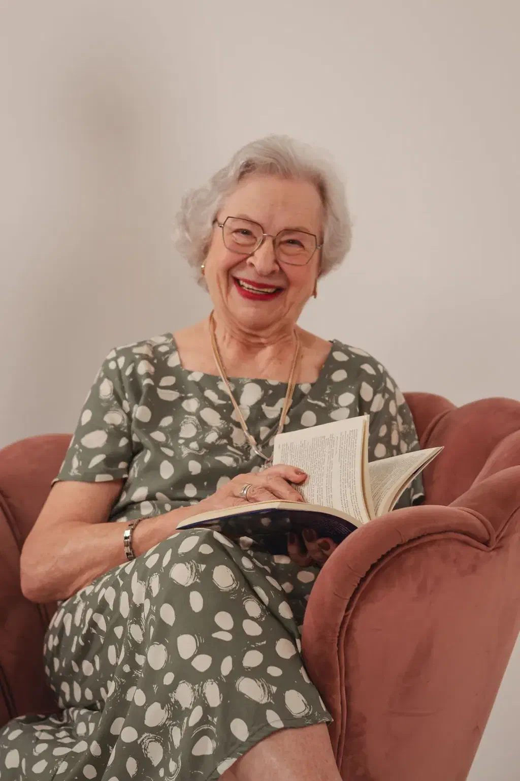 Une femme âgée souriante, un livre ouvert sur les genoux, assise dans un fauteuil