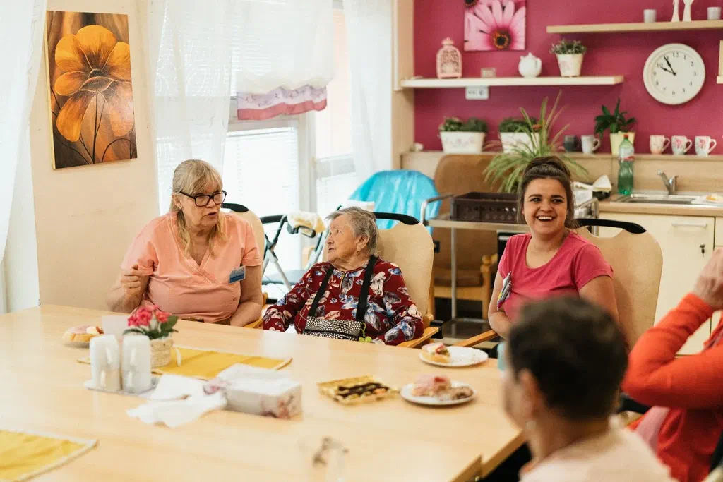 Une femme âgée attablée dans une salle commune lumineuse, entourée d'autres résidentes et de deux accompagnantes