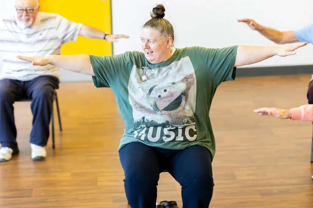 Des personnes âgées assises sur des chaises font un exercice bras écartés dans une salle