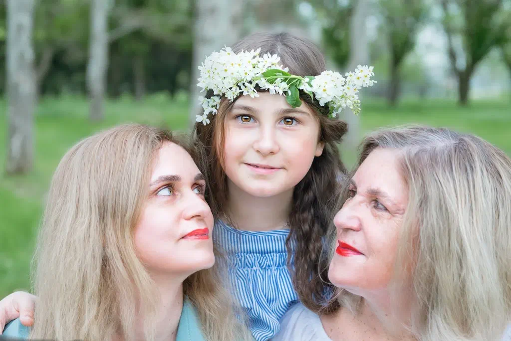 Une fillette couronnée de fleurs blanches entre sa mère et sa grand-mère, dans un parc