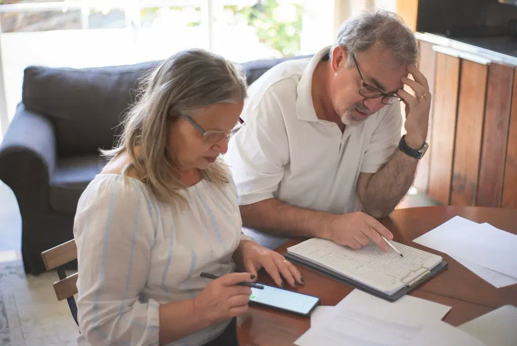 Une femme et un homme aux cheveux gris assis à une table, penchés sur des papiers et un téléphone pour faire des comptes