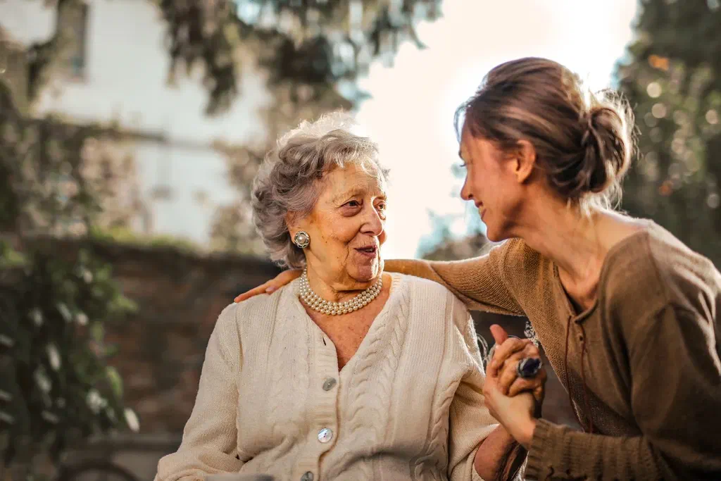 Une femme d'une quarantaine d'années pose la main sur l'épaule de sa mère âgée, assise dans un jardin, et lui tient la main en souriant