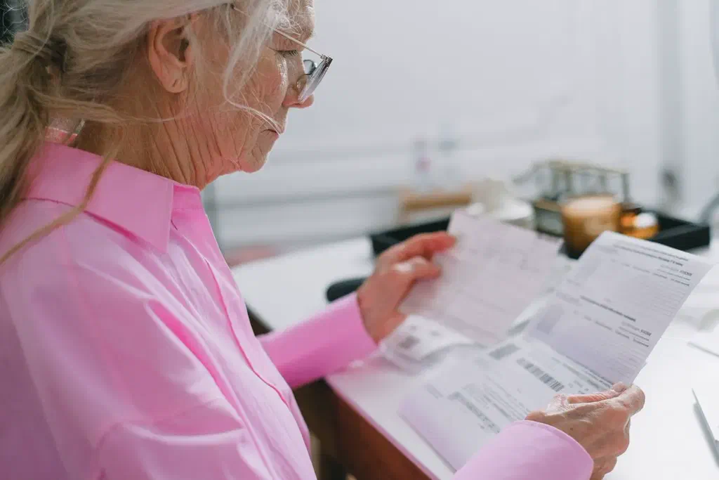 Une femme âgée en chemisier rose, lunettes sur le nez, lit attentivement des documents administratifs à son bureau