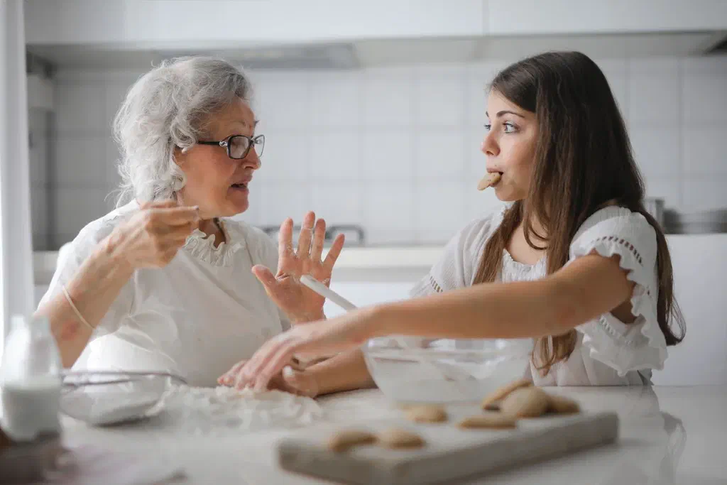 Une femme âgée aux cheveux blancs et une adolescente discutent en préparant des biscuits dans une cuisine claire