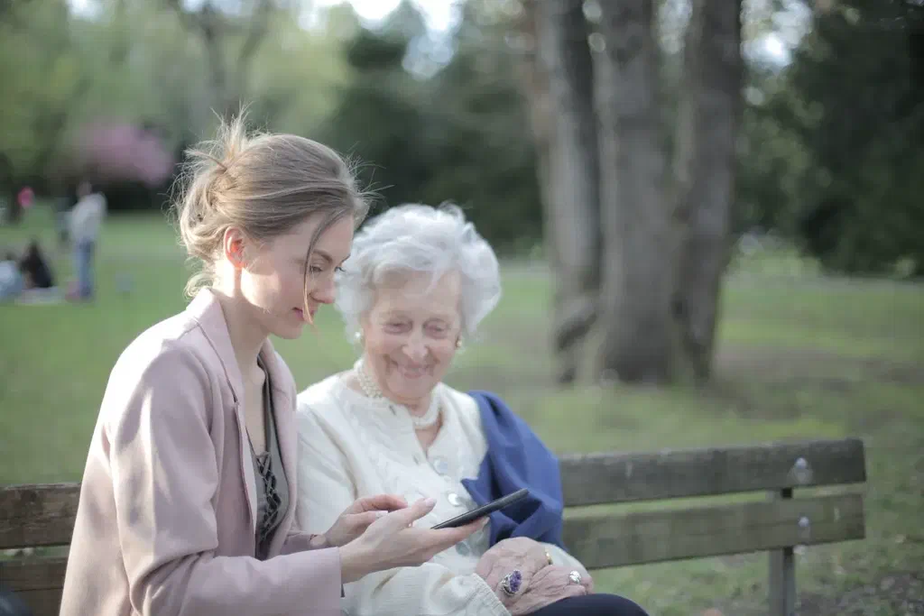 Une jeune femme et une femme âgée assises sur un banc dans un parc, regardant ensemble un téléphone