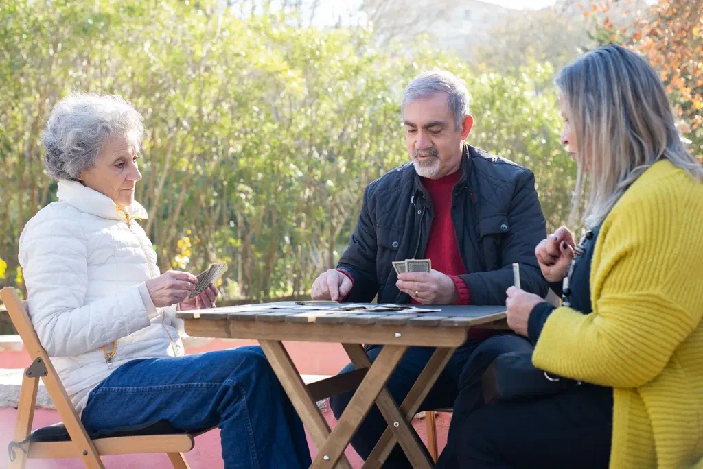 Trois personnes, dont une femme âgée aux cheveux gris, jouent aux cartes autour d'une table en bois dans un jardin ensoleillé