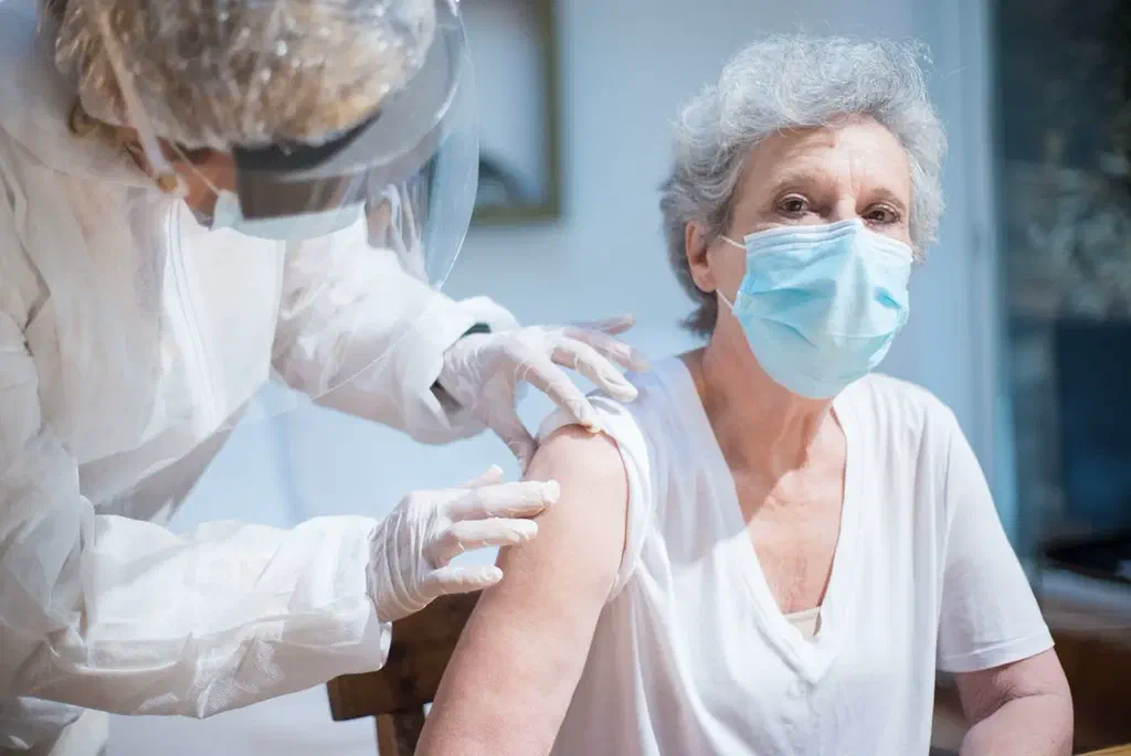 Une femme âgée aux cheveux gris, masquée, regarde l'objectif pendant qu'une soignante en tenue de protection lui prépare le bras pour une injection